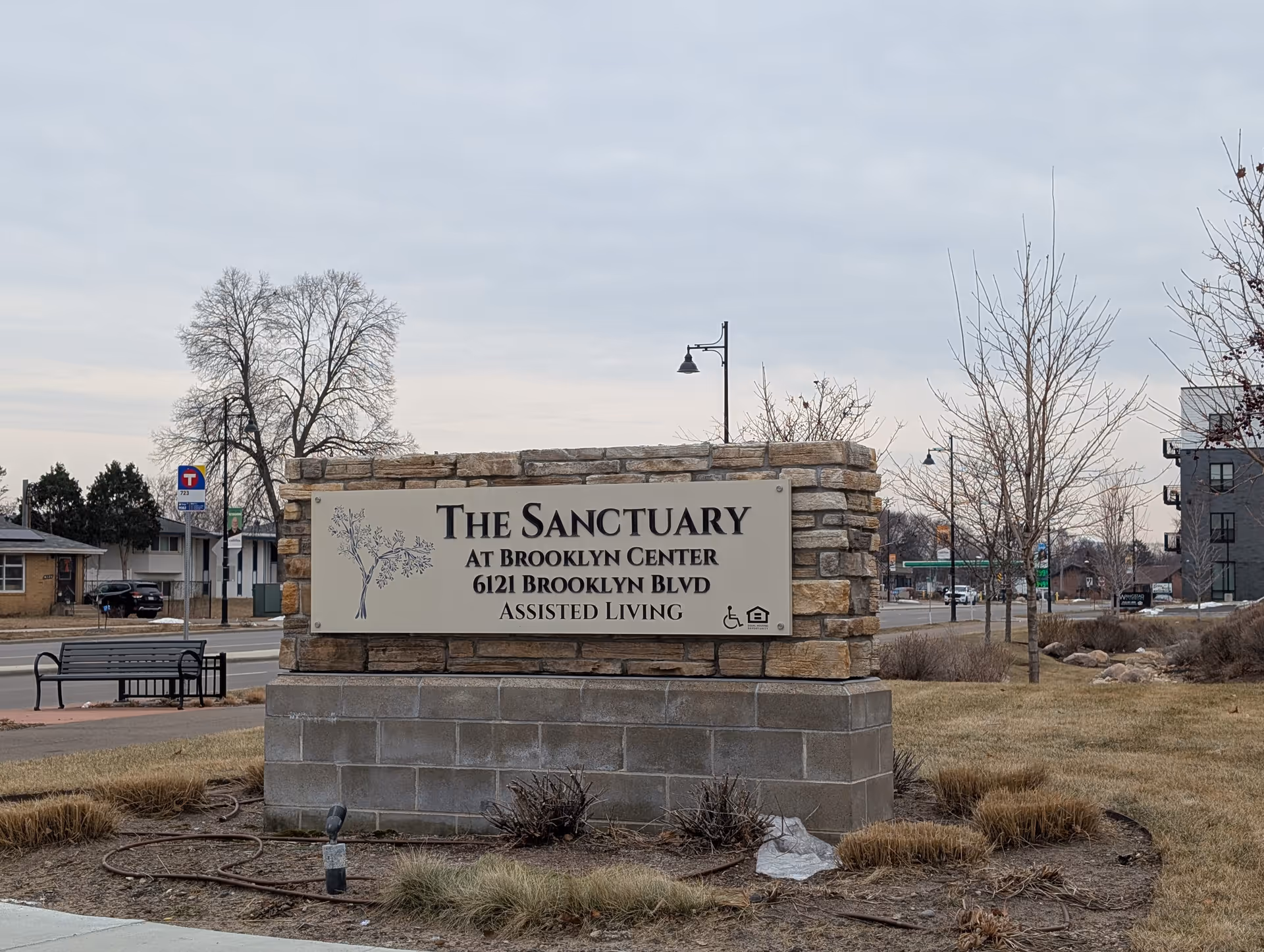 Outdoor stone sign for The Sanctuary at Brooklyn Center, located at 6121 Brooklyn Blvd, indicating it is an assisted living facility. The sign is set on a stone and concrete base with a tree graphic on the left side. Surrounding the sign is a grassy area with some leafless trees and a street with buildings and a bus stop in the background under a cloudy sky.