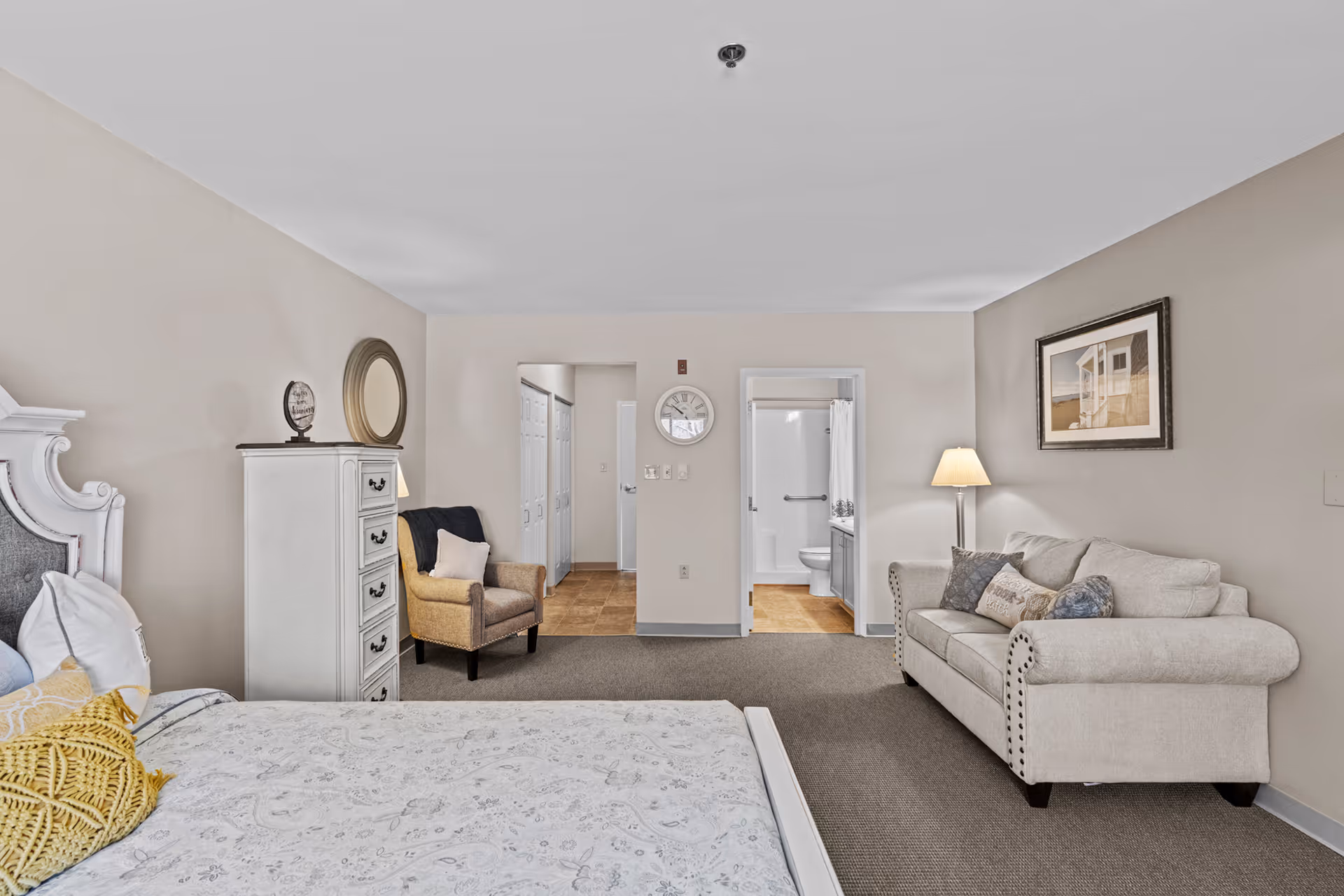 A senior living facility bedroom with a bed on the left side, a white chest of drawers, a beige armchair with a pillow, a beige sofa with multiple cushions, a floor lamp, and a framed picture on the wall. There is a doorway leading to a bathroom with a shower, toilet, and sink visible. The room has beige walls and carpeted flooring.