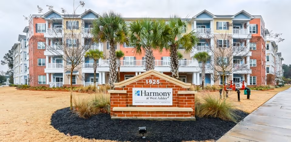 Front exterior view of Harmony at West Ashley, a multi-story senior living facility with balconies, palm trees, and a brick sign displaying the facility name and address number 1925.