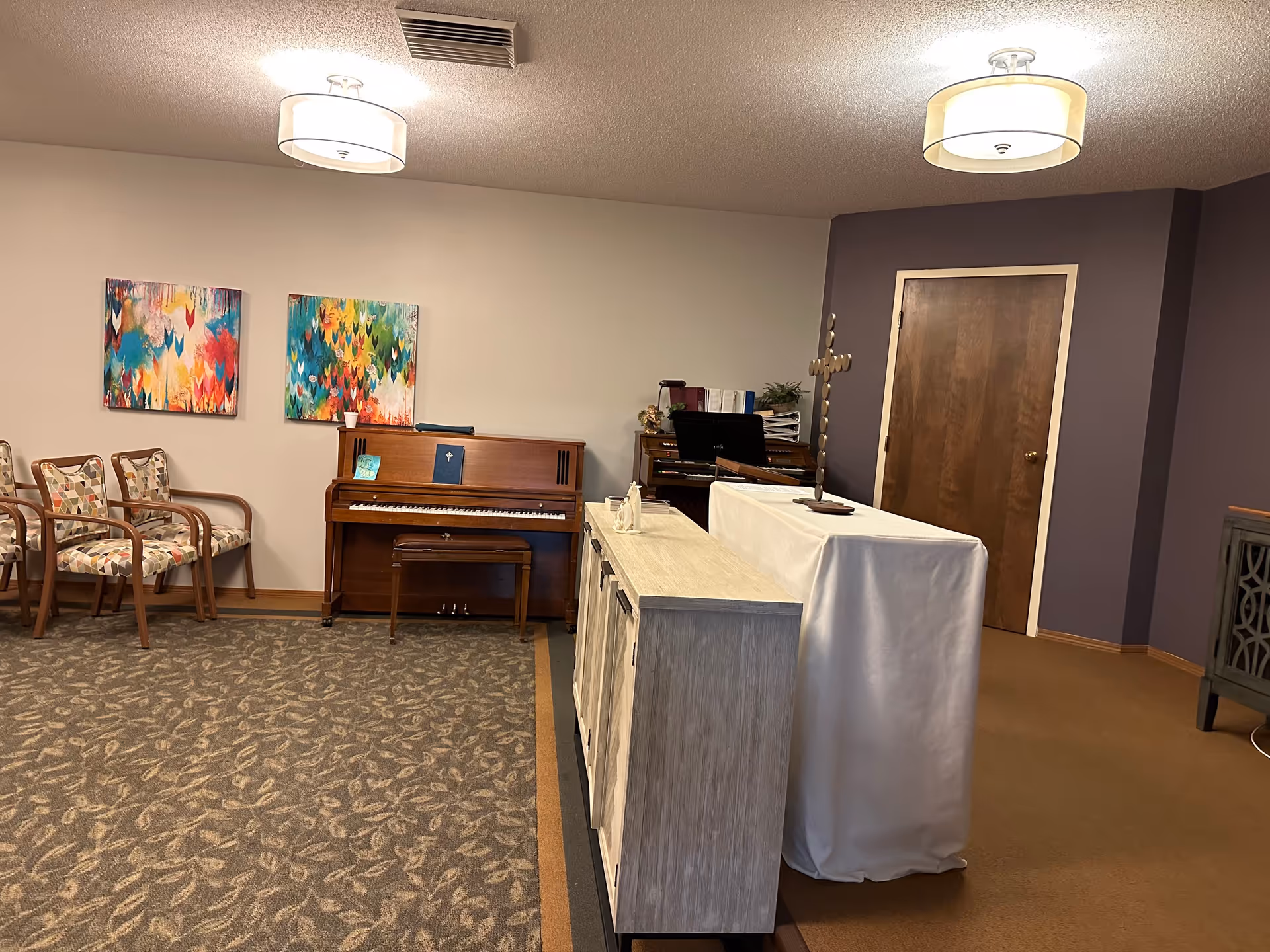 Interior room with a piano against the wall, three patterned chairs, two colorful abstract paintings, a wooden door, and a table covered with a white cloth displaying a decorative cross and figurines. The room has carpeted floors and two ceiling lights.