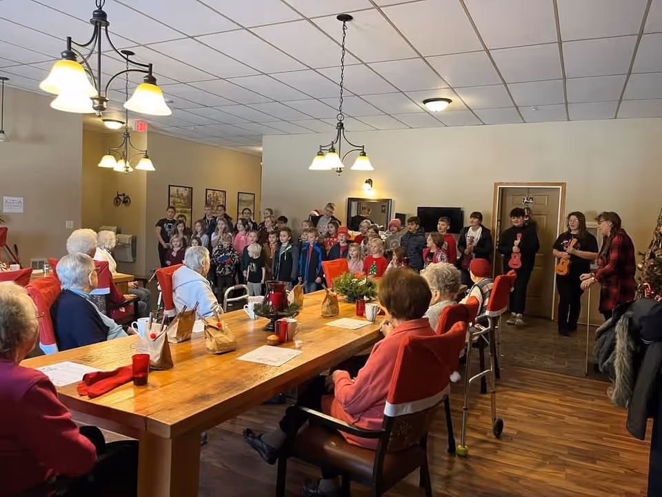 Elderly residents seated around a long dining table watch a group of children performing in a community room.