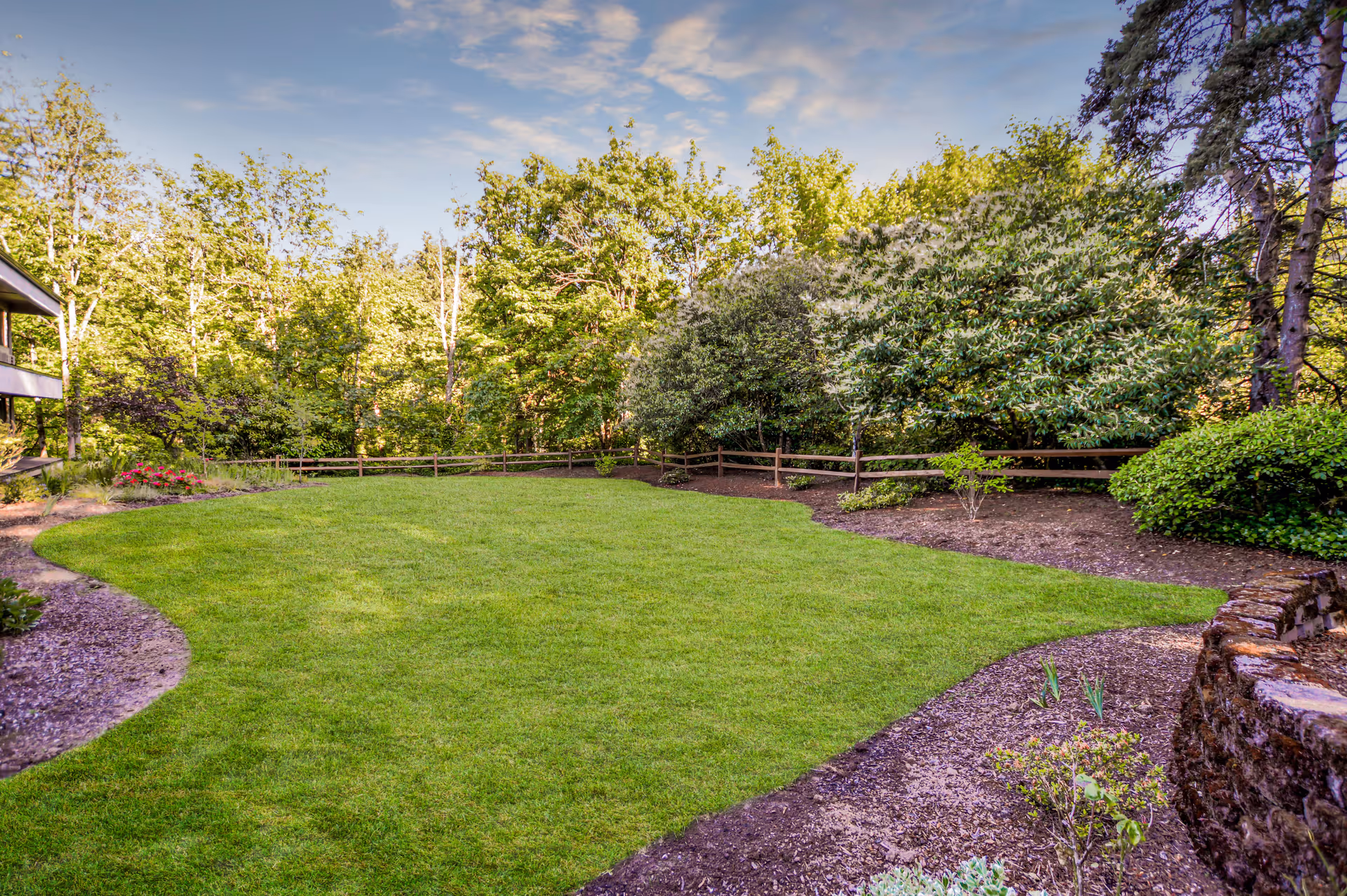 A well-maintained outdoor garden area with a lush green lawn, surrounded by various trees and shrubs. There is a wooden fence along the back and a stone retaining wall on the right side. The sky is clear with some light clouds.