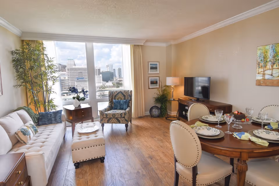 A bright living and dining area in an apartment with large windows showing a cityscape view. The room features a beige sofa with decorative pillows, a patterned armchair, a tufted ottoman, a wooden dining table set with plates, glasses, and napkins, a TV on a wooden stand, and various plants and wall art.
