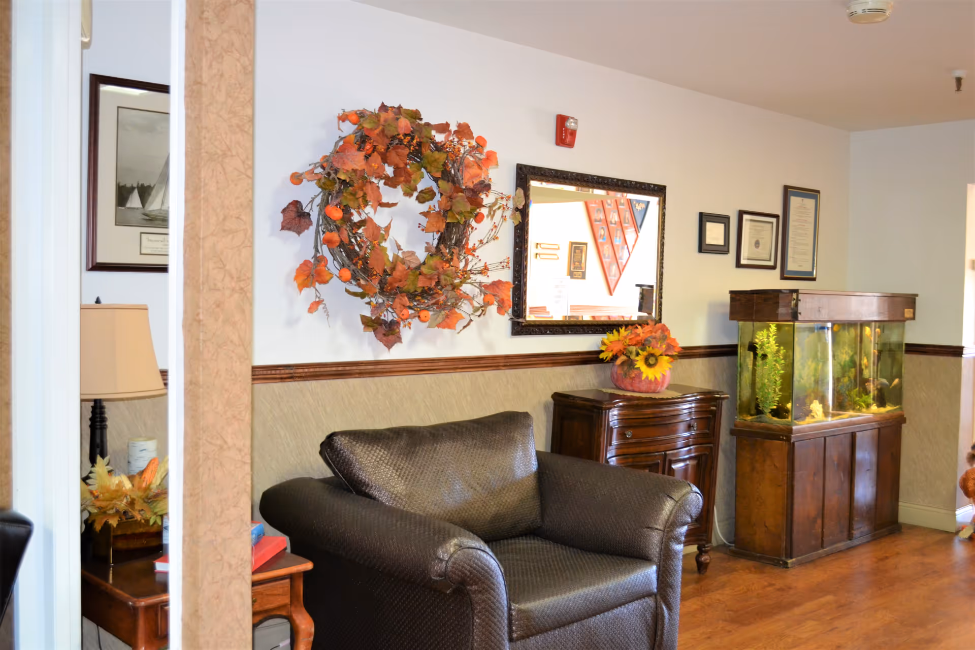 Cozy lounge area with a leather armchair, wooden side tables, a fall wreath and mirror on the wall, and a large fish tank.