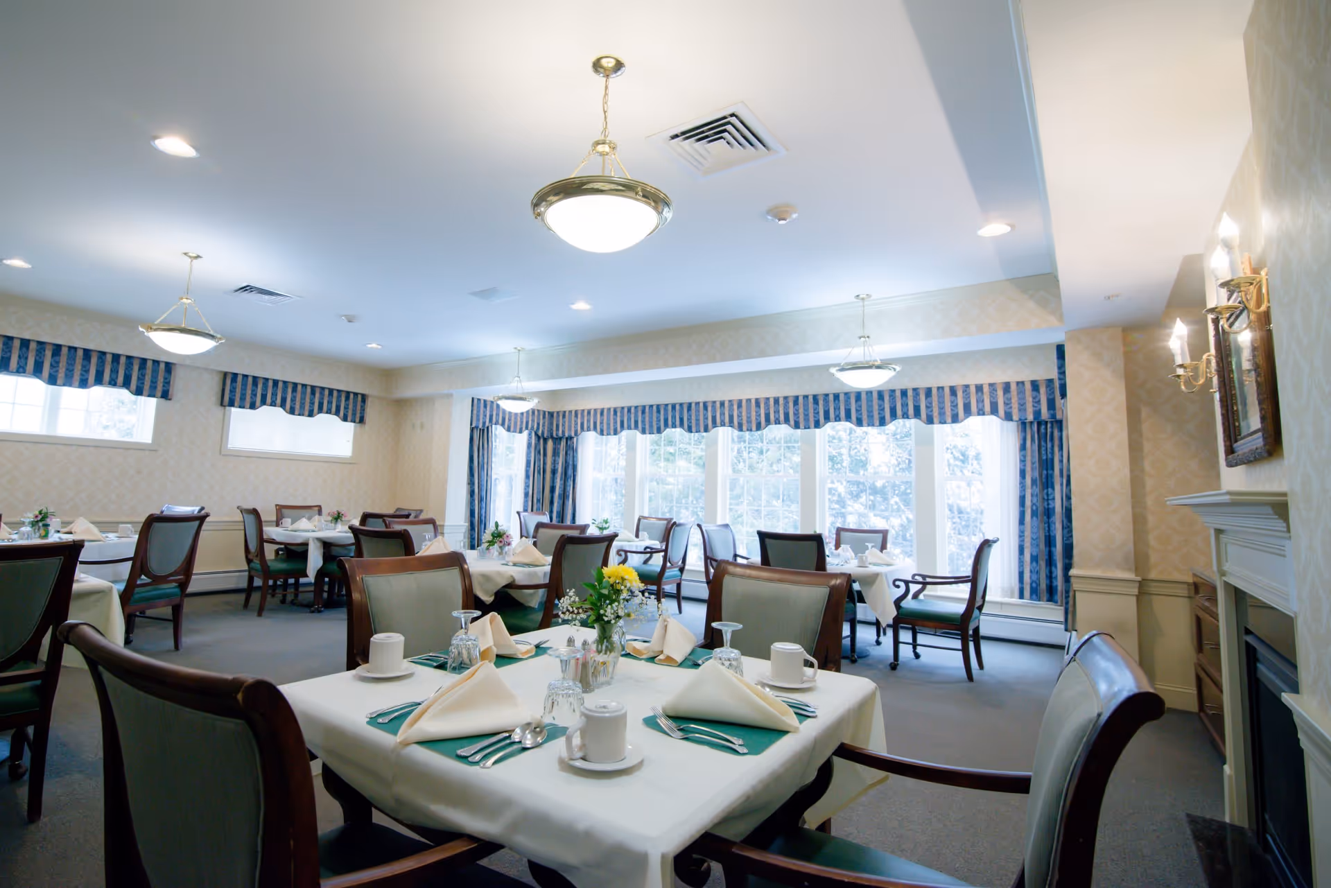 A bright dining room with multiple tables set with white tablecloths, folded napkins, cups, glasses, and silverware. The room features large windows with blue and white striped curtains, elegant hanging light fixtures, and a fireplace on the right wall.