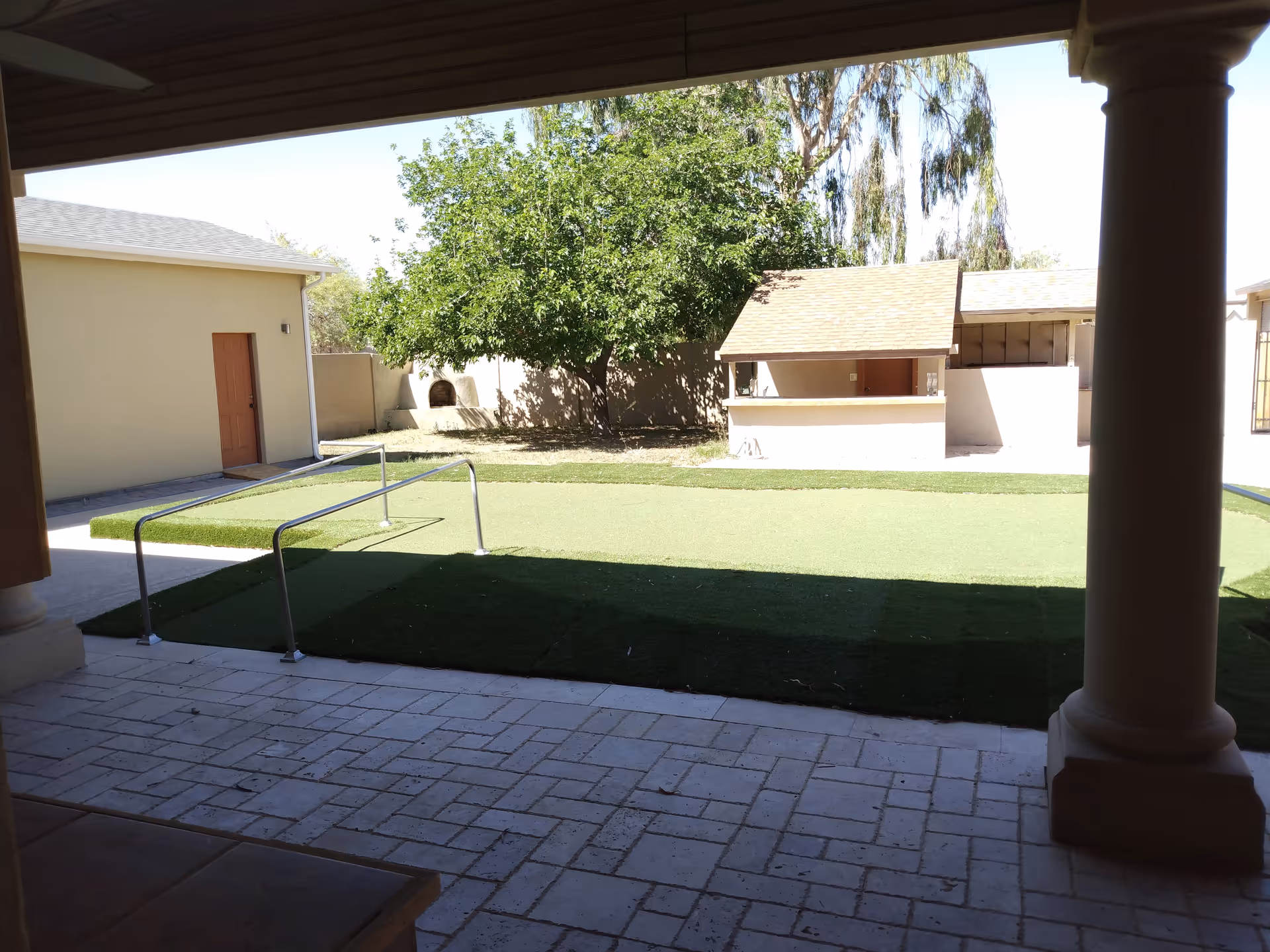 View of an outdoor courtyard area at Biltmore Care Home 2 with artificial grass, a metal handrail, a tree, a small structure with a roof, and a beige building with a brown door. The area is partially shaded by a covered patio with columns.