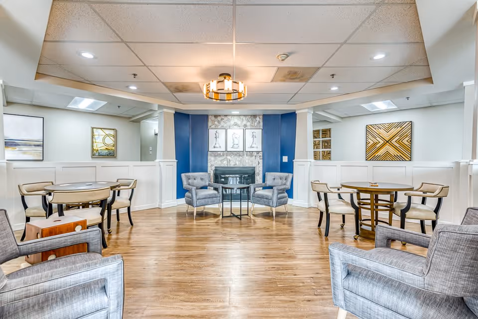 A bright and spacious senior living common area with wooden flooring, several upholstered armchairs, and round tables with chairs. The room features a central fireplace with a blue accent wall and three framed chess piece artworks above it. Modern ceiling lights illuminate the space, and abstract paintings decorate the white walls.