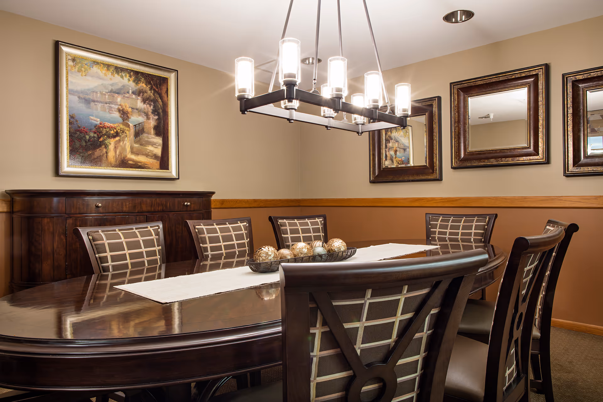 A dining room with a large dark wooden table surrounded by eight chairs with patterned upholstery. A modern chandelier with multiple lights hangs above the table. On the wall, there are three framed mirrors and a framed painting depicting a scenic outdoor view with water and greenery. A dark wooden sideboard is positioned against the wall beneath the painting.
