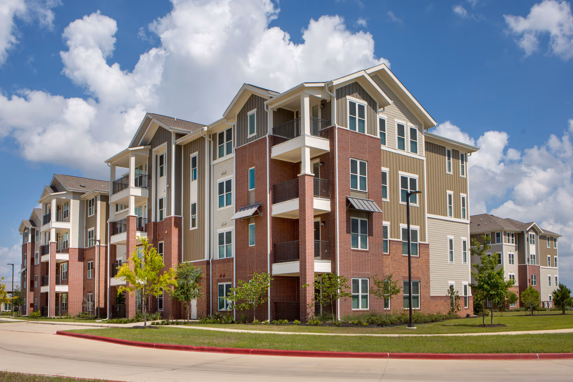Exterior view of a modern multi-story senior living facility building with brick and beige siding, balconies, and a well-maintained lawn under a partly cloudy sky.
