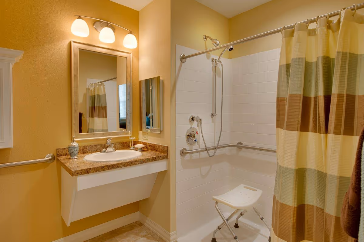 A bathroom with a wall-mounted sink and a large mirror above it. The shower area has white tiled walls, a grab bar, a handheld showerhead, and a white shower chair. The shower curtain has horizontal stripes in shades of green, brown, and beige. The walls are painted yellow, and there is a three-light fixture above the mirror.