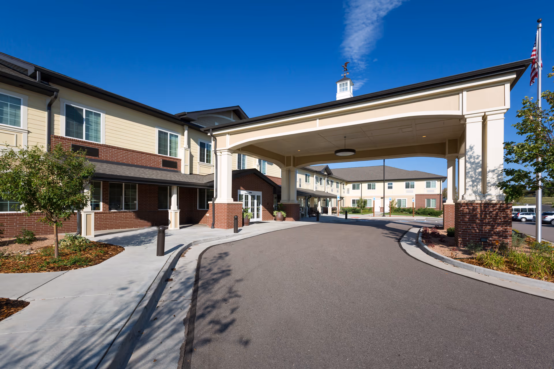Exterior view of Peakview Assisted Living and Memory Care facility showing a covered entrance with a driveway, landscaped areas with small trees and shrubs, and a two-story building with beige siding and brick accents under a clear blue sky.