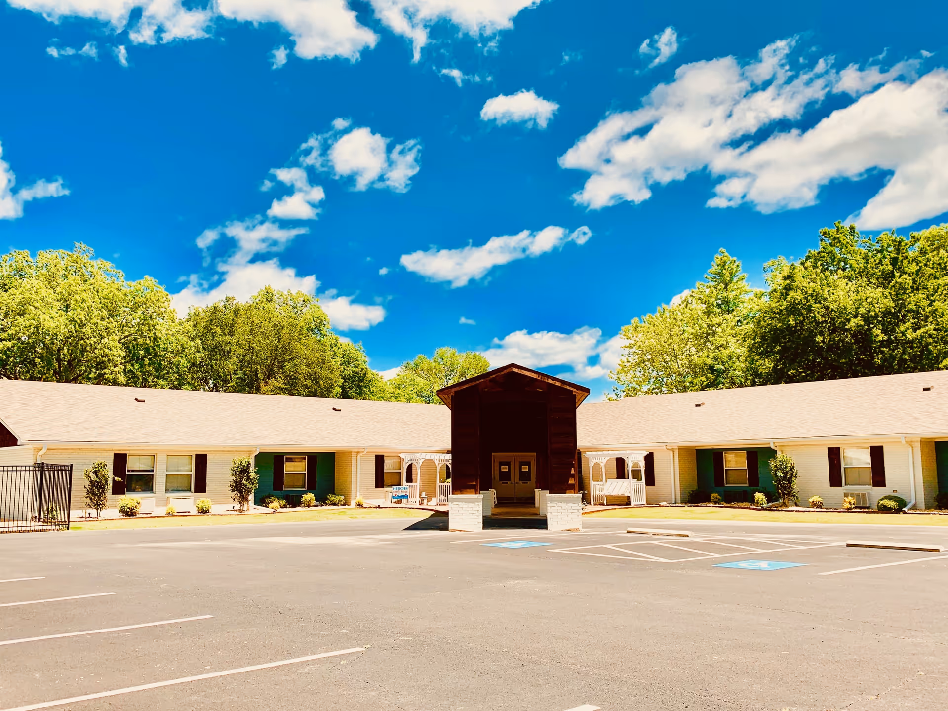 Exterior view of a single-story building with a light-colored roof and walls, surrounded by green trees under a blue sky with scattered clouds. The building has a central covered entrance with double doors and a paved parking lot in front, including marked handicap parking spaces.