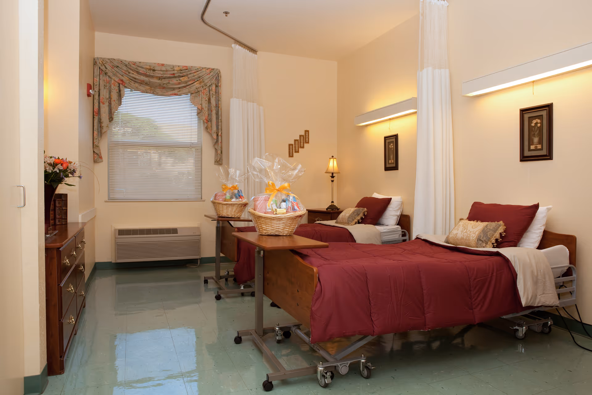 A room with two hospital-style beds, each made up with burgundy bedspreads, pillows, and beige blankets. Each bed has a small overbed table with a gift basket wrapped in cellophane and tied with a yellow ribbon. The room has a window with floral valance curtains, a wooden dresser with flowers on top, a lamp on a nightstand between the beds, and framed artwork on the walls. The floor is a shiny green linoleum.
