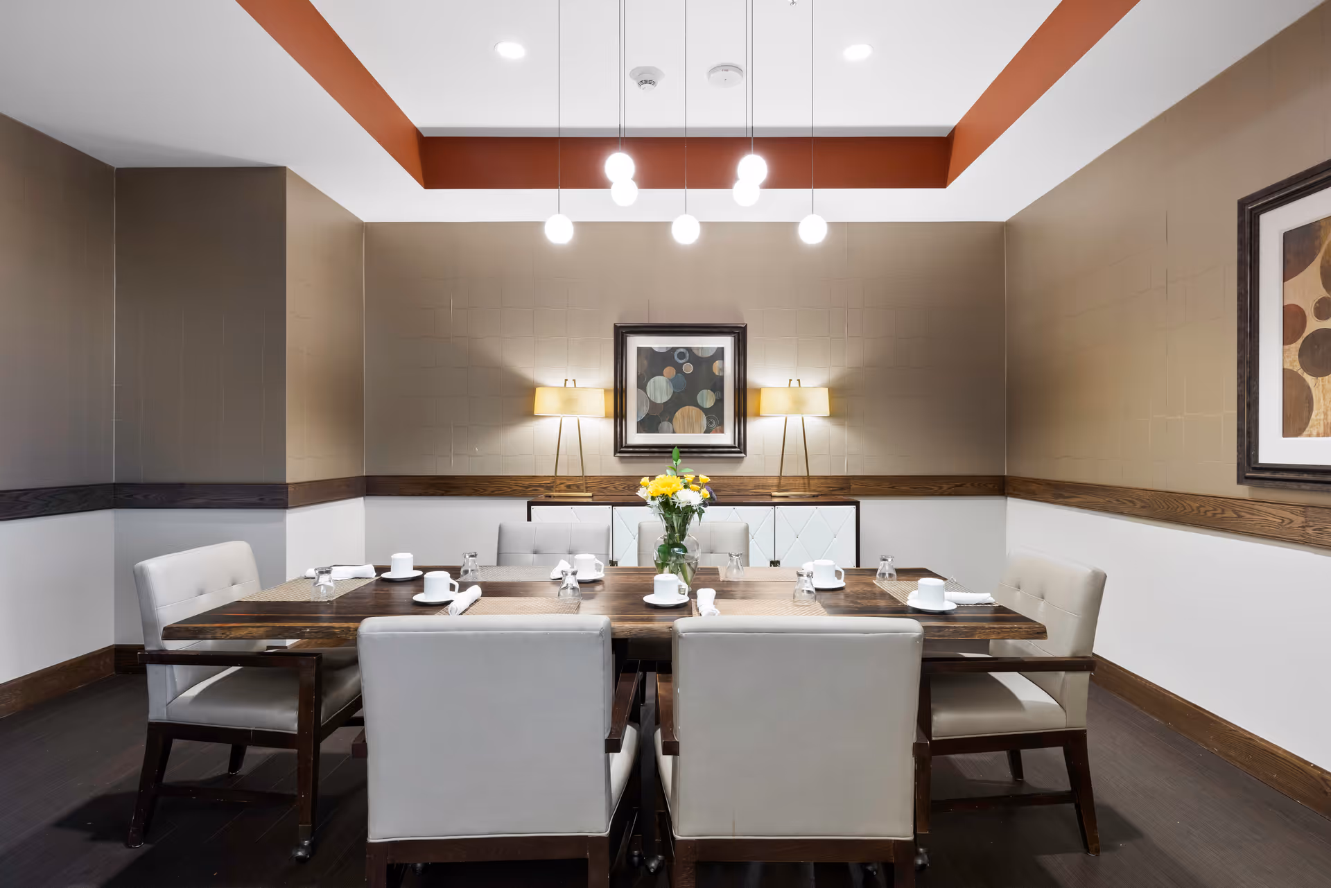 A dining room with a wooden table set for six people with white cups, napkins, and glasses. The room has beige and white walls with wooden trim, two standing lamps, a framed abstract painting, and a ceiling with hanging pendant lights. A vase with yellow flowers is placed in the center of the table.