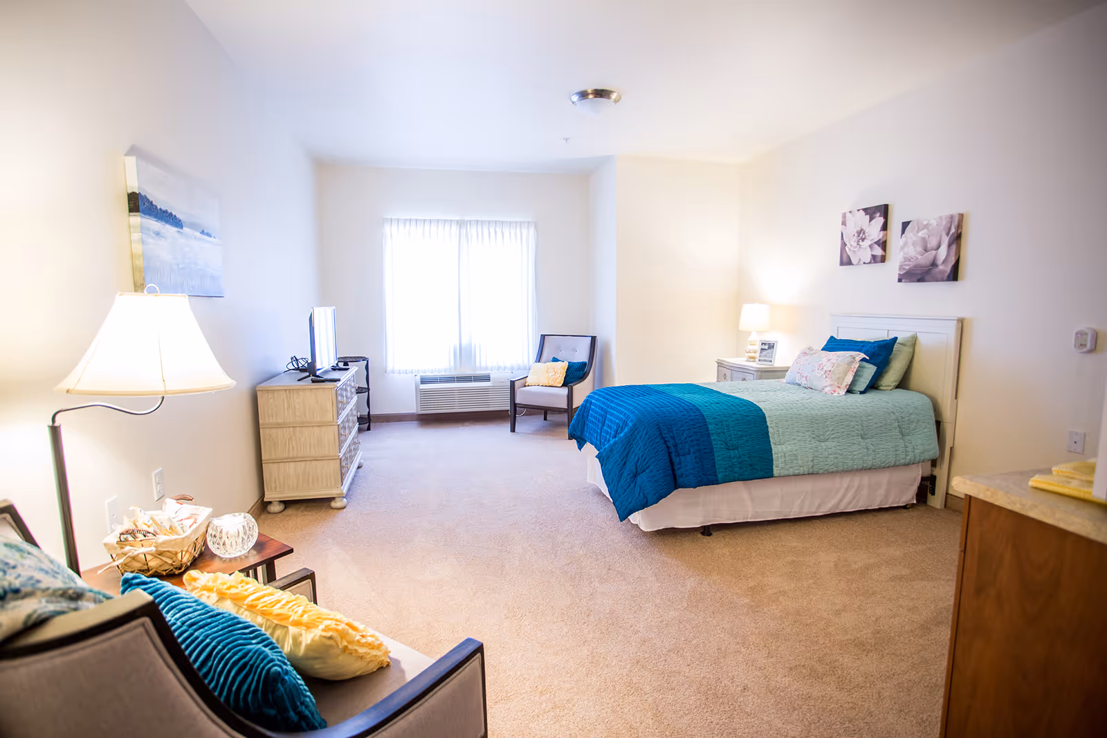 A bright and cozy senior living bedroom with a single bed covered in blue and teal bedding, a nightstand with a lamp, a dresser with a TV on top, a cushioned chair with pillows near a window with white curtains, and a small sofa with colorful pillows. The walls are decorated with floral artwork.