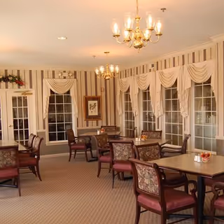 A dining room with multiple tables and chairs arranged neatly. The room features striped wallpaper, large windows with draped curtains, and two chandeliers hanging from the ceiling. The tables have small decorative items on them.