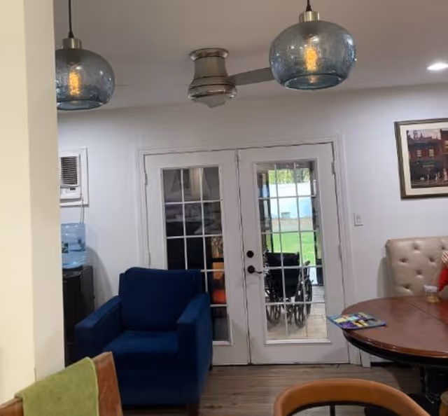 Interior view of a living room area with a blue armchair, a round wooden table with chairs, and a beige cushioned bench. There are two glass pendant lights hanging from the ceiling and a ceiling fan. French doors with glass panes lead outside to a patio where a wheelchair is visible. A framed picture hangs on the wall.