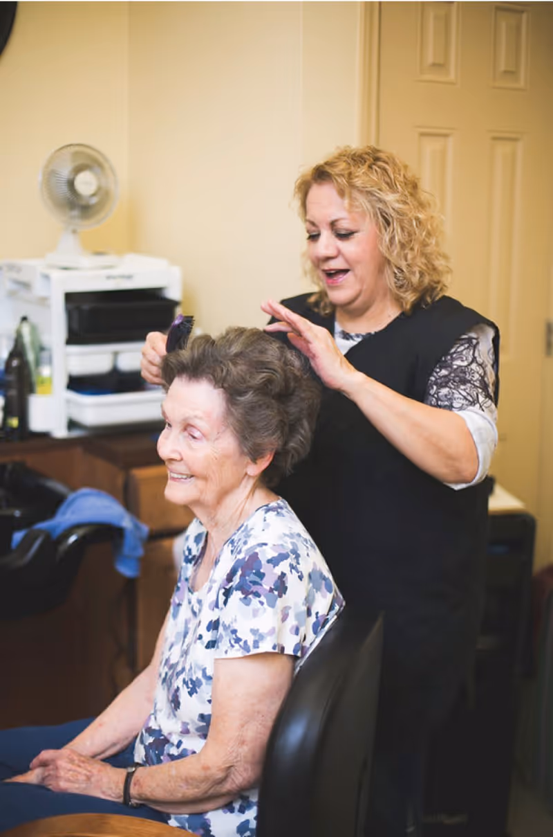 An elderly woman with short curly hair sits in a chair smiling while a hairstylist stands behind her, combing and styling her hair in a cozy indoor setting.