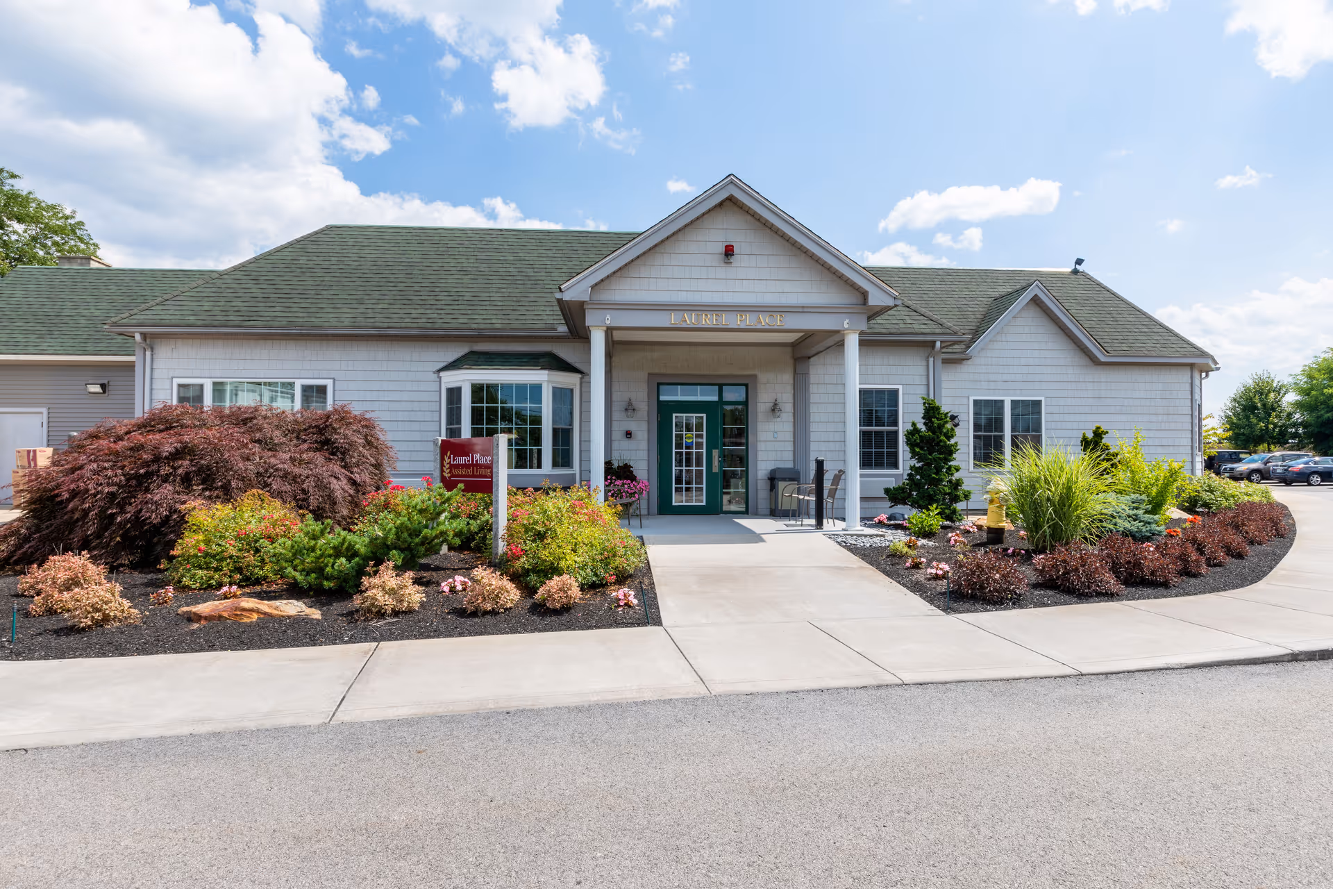 Front exterior view of Laurel Place Assisted Living building with a green roof, white siding, and a covered entrance. The entrance is flanked by landscaped bushes and flowers, with a clear blue sky and some clouds overhead.