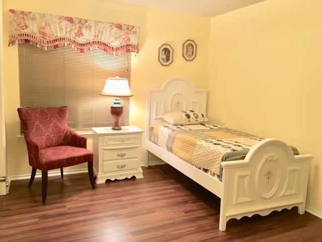 A yellow-painted bedroom with an ornate white bed, nightstand, lamp, pink chair, and window blinds.