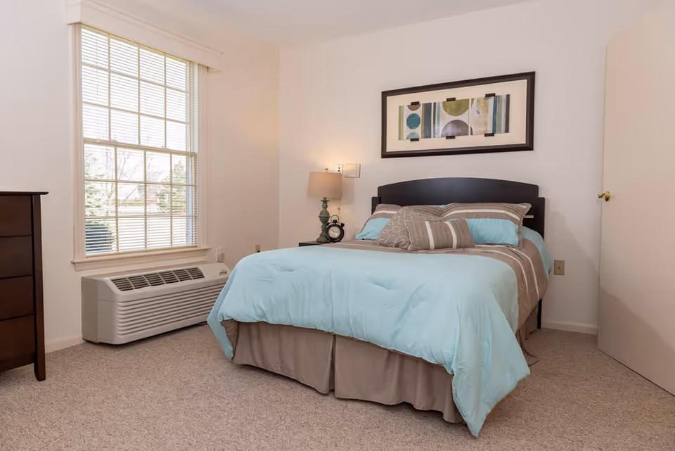 Well-lit bedroom with a double bed dressed in teal and brown bedding, a nightstand with a lamp, and a large window.