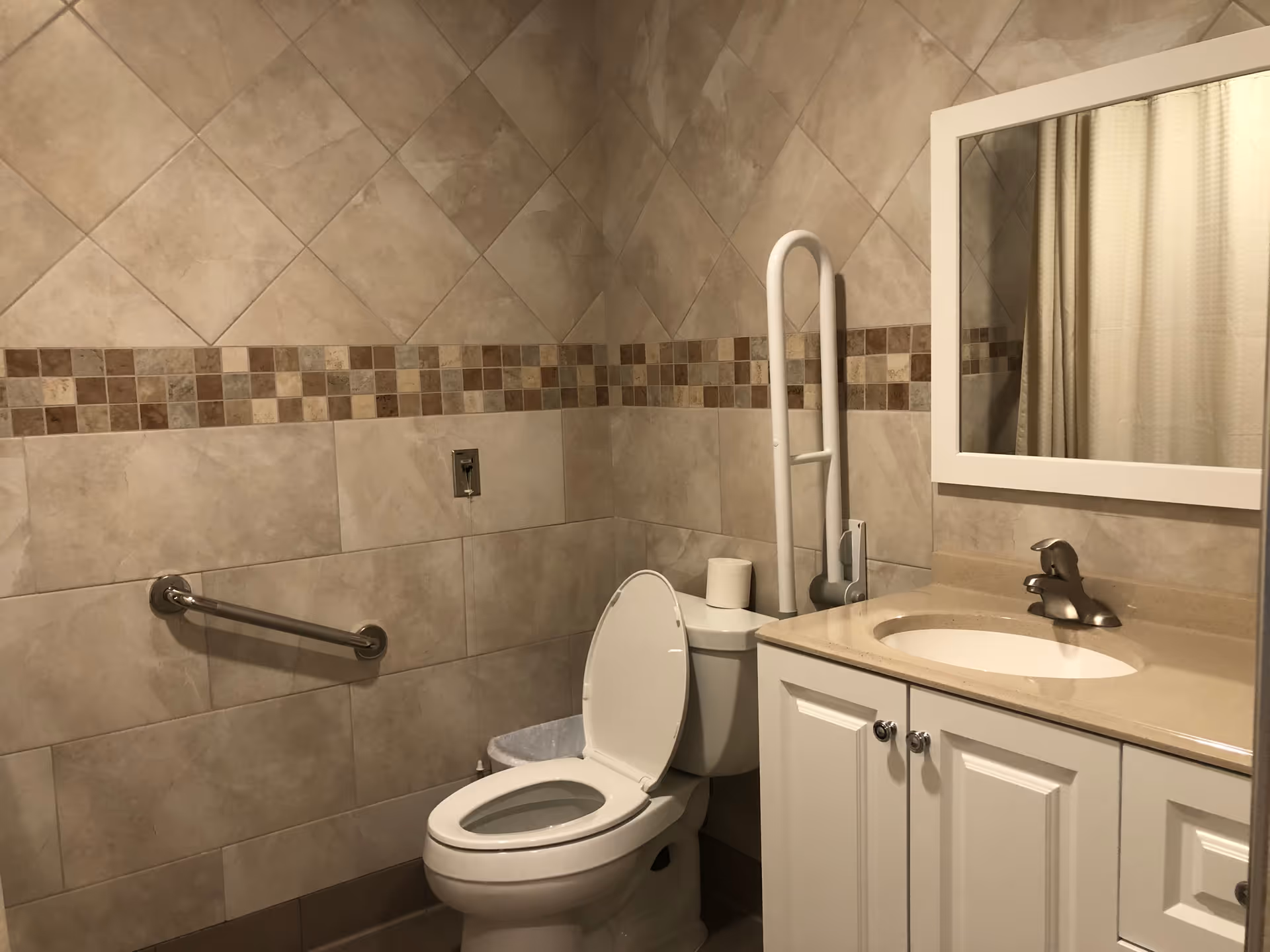 A clean bathroom with beige tiled walls featuring a decorative strip of small square tiles. The bathroom includes a white toilet with the lid open, a grab bar on the wall beside it, a white vanity with a beige countertop and an oval sink, a silver faucet, and a large rectangular mirror above the sink. There is also a white vertical grab bar next to the toilet and a roll of toilet paper on top of the toilet tank.