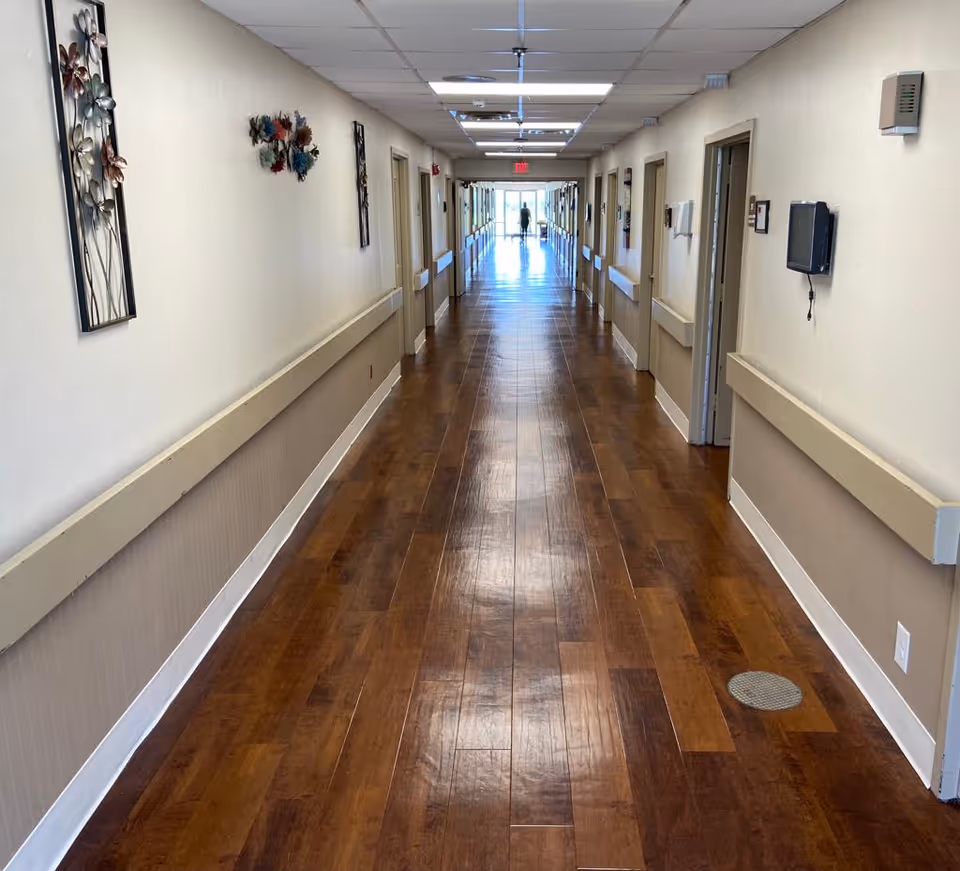 Long, polished wood-floored hallway in a healthcare facility with handrails and doors lining both sides.