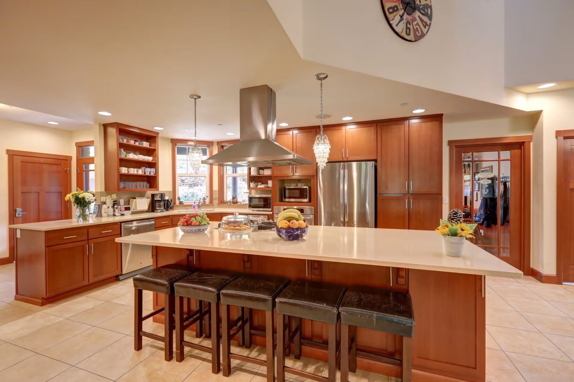 A spacious kitchen with a large island countertop featuring four black cushioned stools. The kitchen has wooden cabinets, stainless steel appliances including a refrigerator and built-in microwave, and two pendant lights hanging above the island. There are bowls of fruit and a plate of cookies on the island. The room has tiled floors, a large clock on the wall, and windows letting in natural light.