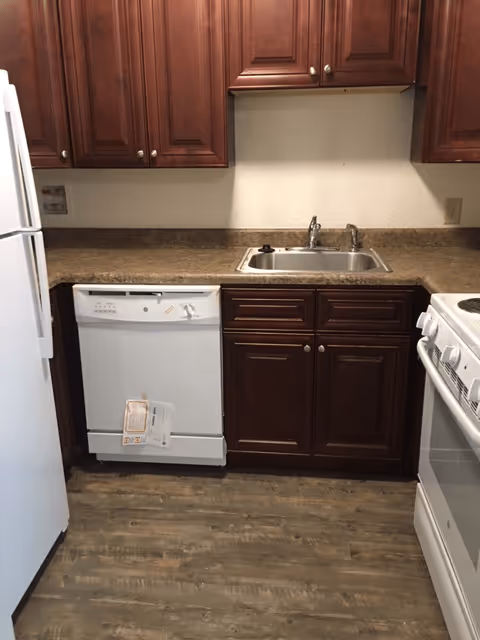 Small kitchen area with dark wooden cabinets, a white refrigerator on the left, a white dishwasher below the counter, a stainless steel sink with a faucet, and a white stove on the right. The countertop is brown, and the floor has a wood-like finish.