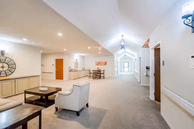 A bright and spacious hallway in a senior living facility with beige walls and carpet. The hallway features a seating area with two armchairs and a coffee table on the left, a large decorative wall clock, a small dining table with chairs further down, and several doors along the corridor. The ceiling has recessed lighting and hanging lantern-style light fixtures, with a door visible at the far end of the hallway.