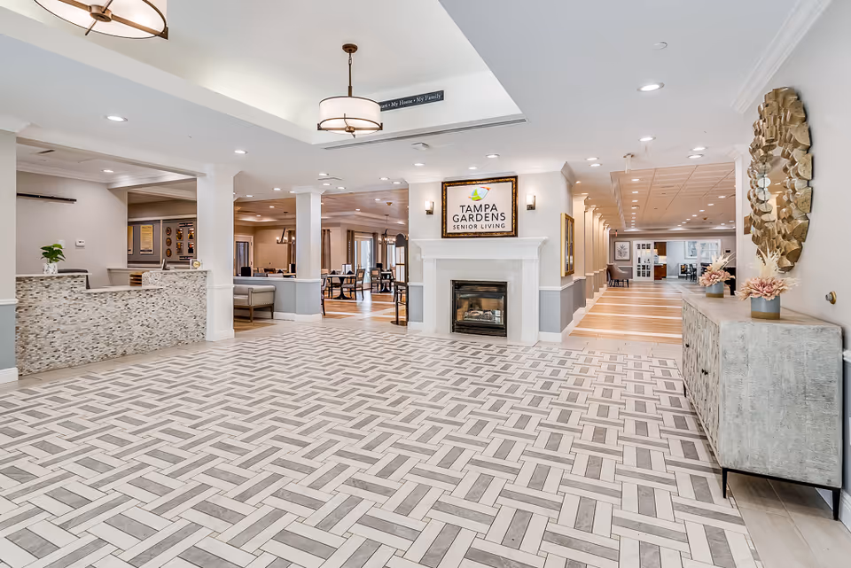 Spacious and well-lit lobby area of Tampa Gardens Senior Living featuring a patterned tile floor, a reception desk on the left, a fireplace with a framed Tampa Gardens Senior Living sign above it in the center, and a decorative console table with flowers and a large round mirror on the right. The background shows a hallway and a dining area with tables and chairs.