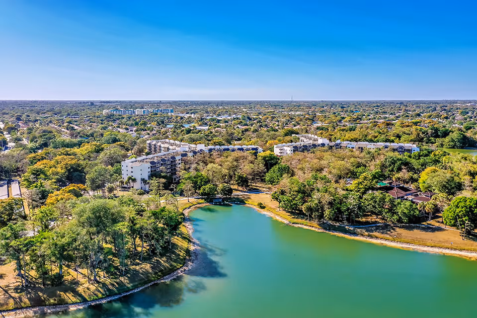Aerial view of a senior living facility named Forest Trace Senior Living surrounded by lush green trees and a large body of water in the foreground under a clear blue sky.