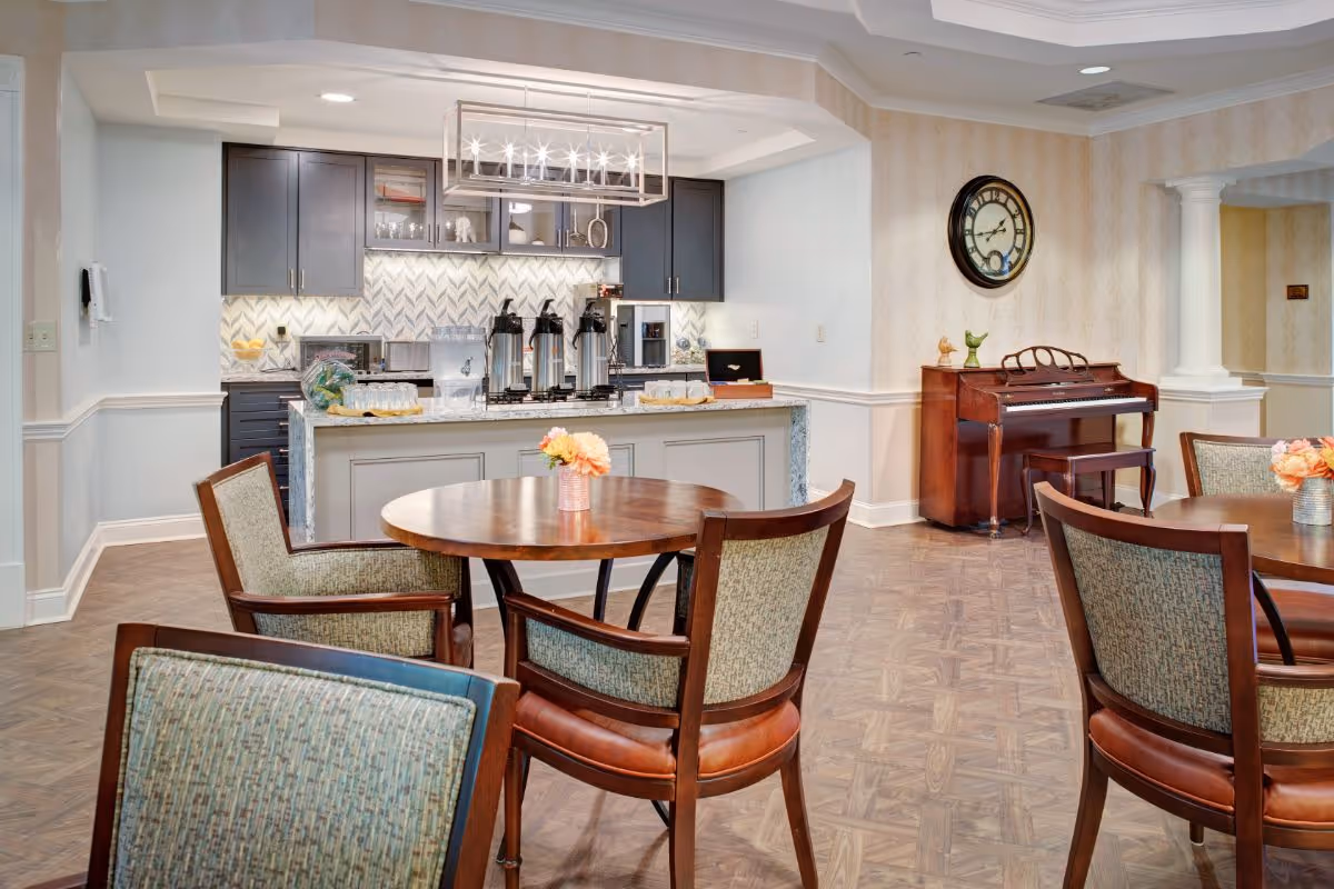 A bright dining area with round wooden tables and upholstered chairs, a service counter and kitchenette in the background and a piano against the wall.