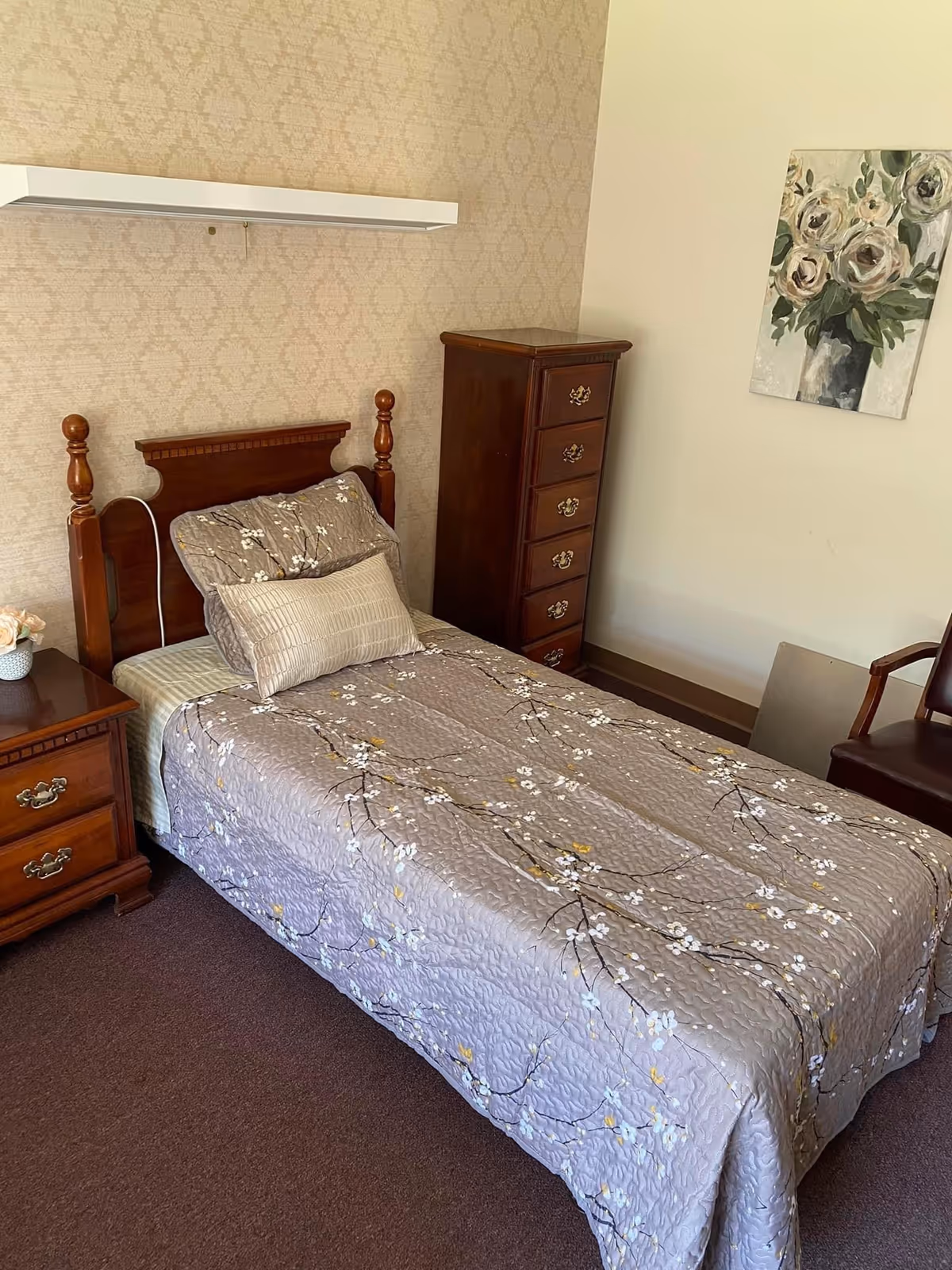 A small bedroom with a single wooden bed covered with a gray floral-patterned quilt and matching pillows. Next to the bed is a wooden nightstand with two drawers and a small flower pot on top. Against the wall is a tall wooden chest of drawers. A painting of white flowers in a vase hangs on the wall above a brown chair. The walls have a light beige patterned wallpaper and the floor is carpeted in brown.