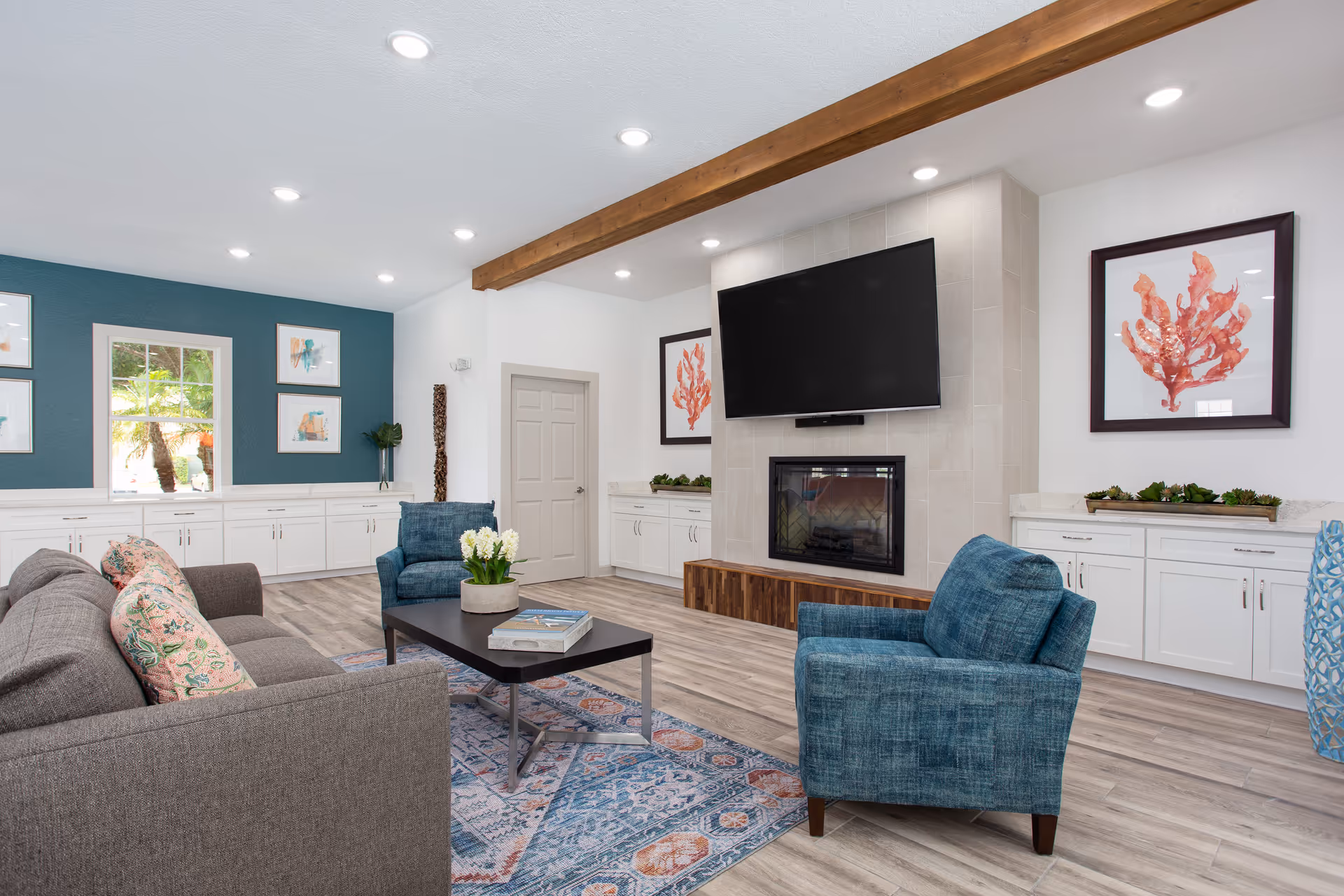 A bright and modern living room with a gray sofa adorned with patterned pillows, two blue armchairs, a black coffee table with a plant and books, and a large flat-screen TV mounted above a fireplace. The room features light wood flooring, white cabinetry along the walls, decorative framed coral artwork, a window with a view of greenery outside, and recessed ceiling lights with a wooden beam accent.