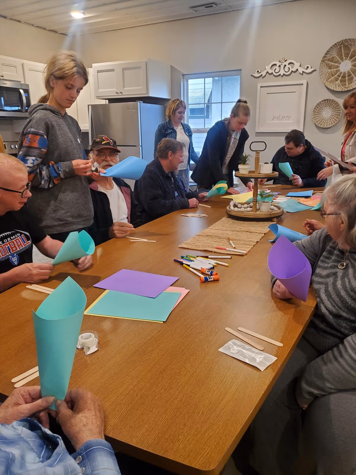 A group of elderly people and caregivers gathered around a large wooden table in a kitchen or dining area, engaging in a craft activity with colored paper, markers, glue sticks, and popsicle sticks. The room has white cabinets, a refrigerator, and decorative wall hangings.