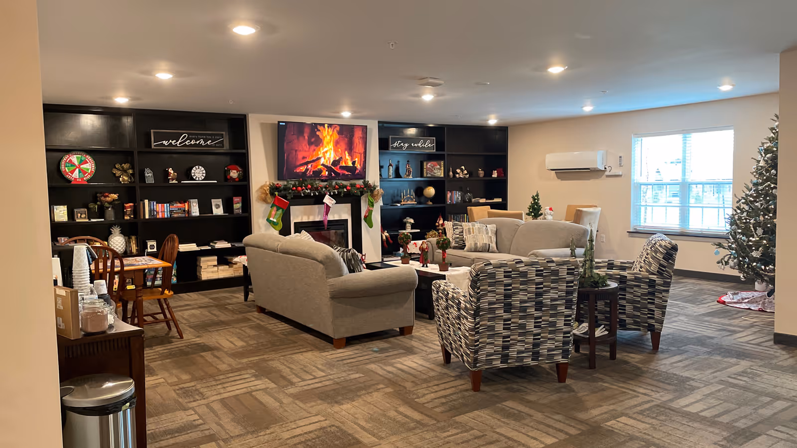 A cozy living room area in a retirement community decorated for the holidays. The room features two gray sofas, two patterned armchairs, a coffee table with holiday decorations, and a Christmas tree near a window. A fireplace with a digital fire display is centered between two black bookshelves filled with books and decorative items. The room is well-lit with ceiling lights and has a carpeted floor.