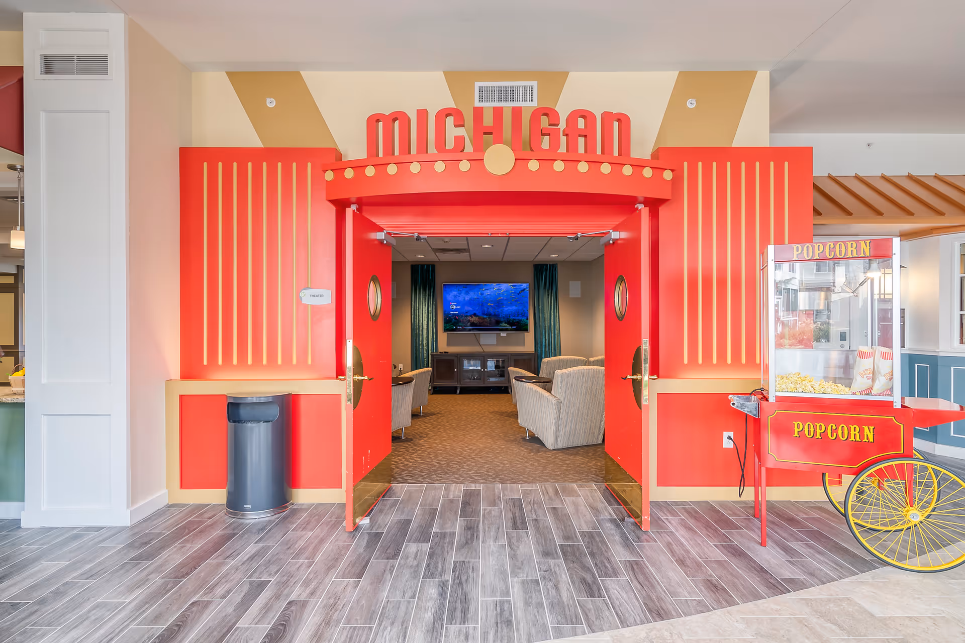 Bright red 'Michigan' themed theater entrance with double doors opening to a seating area and TV, flanked by a popcorn cart.