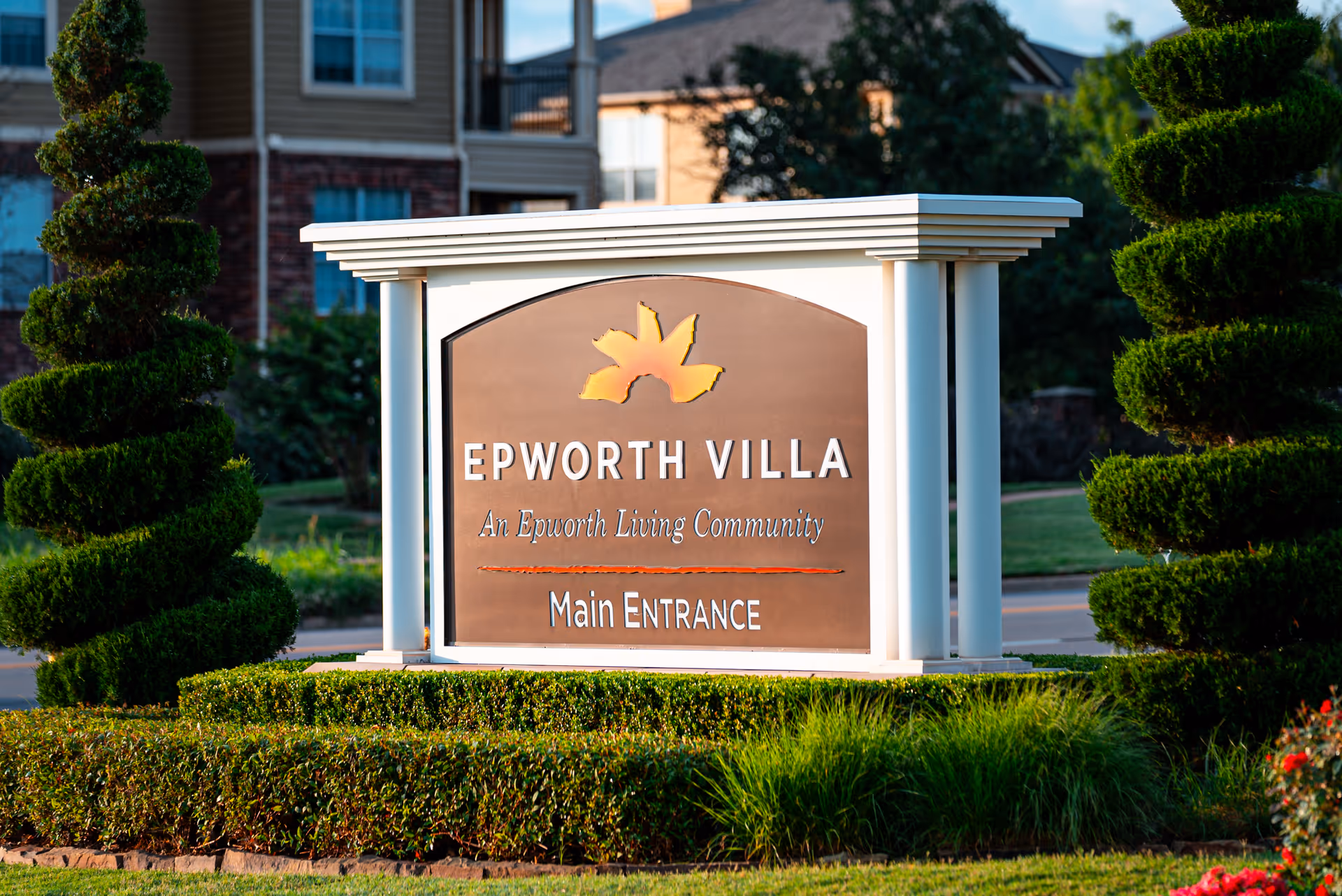 A large sign for Epworth Villa, an Epworth Living Community, indicating the main entrance. The sign is surrounded by neatly trimmed bushes and spiral-shaped topiary trees, with residential buildings visible in the background.