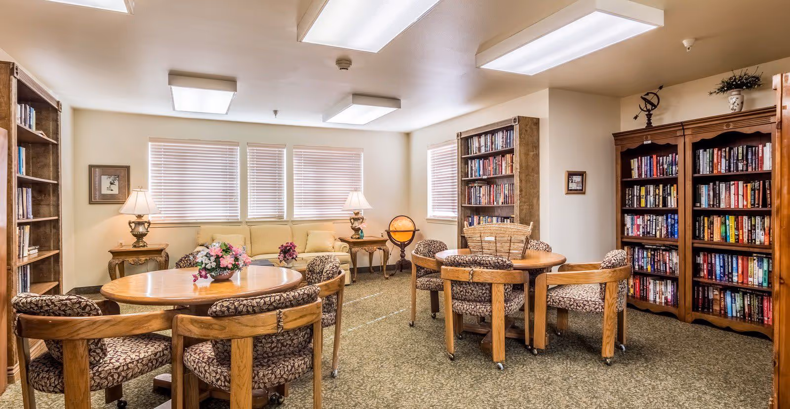 Cozy communal library lounge with round wooden tables, upholstered chairs, bookshelves and a sofa beneath three windows.