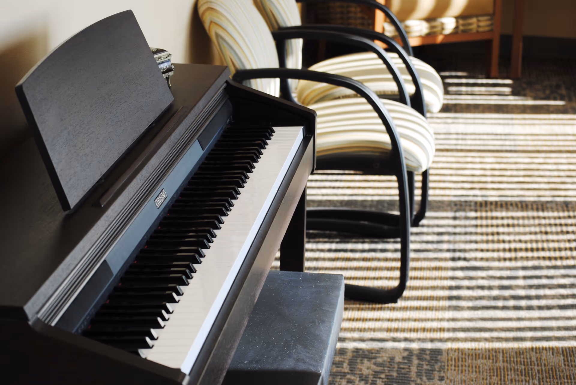 A black digital piano with a matching bench is positioned near two striped cushioned chairs with black armrests. Sunlight streams through a window, casting striped shadows on the carpeted floor.