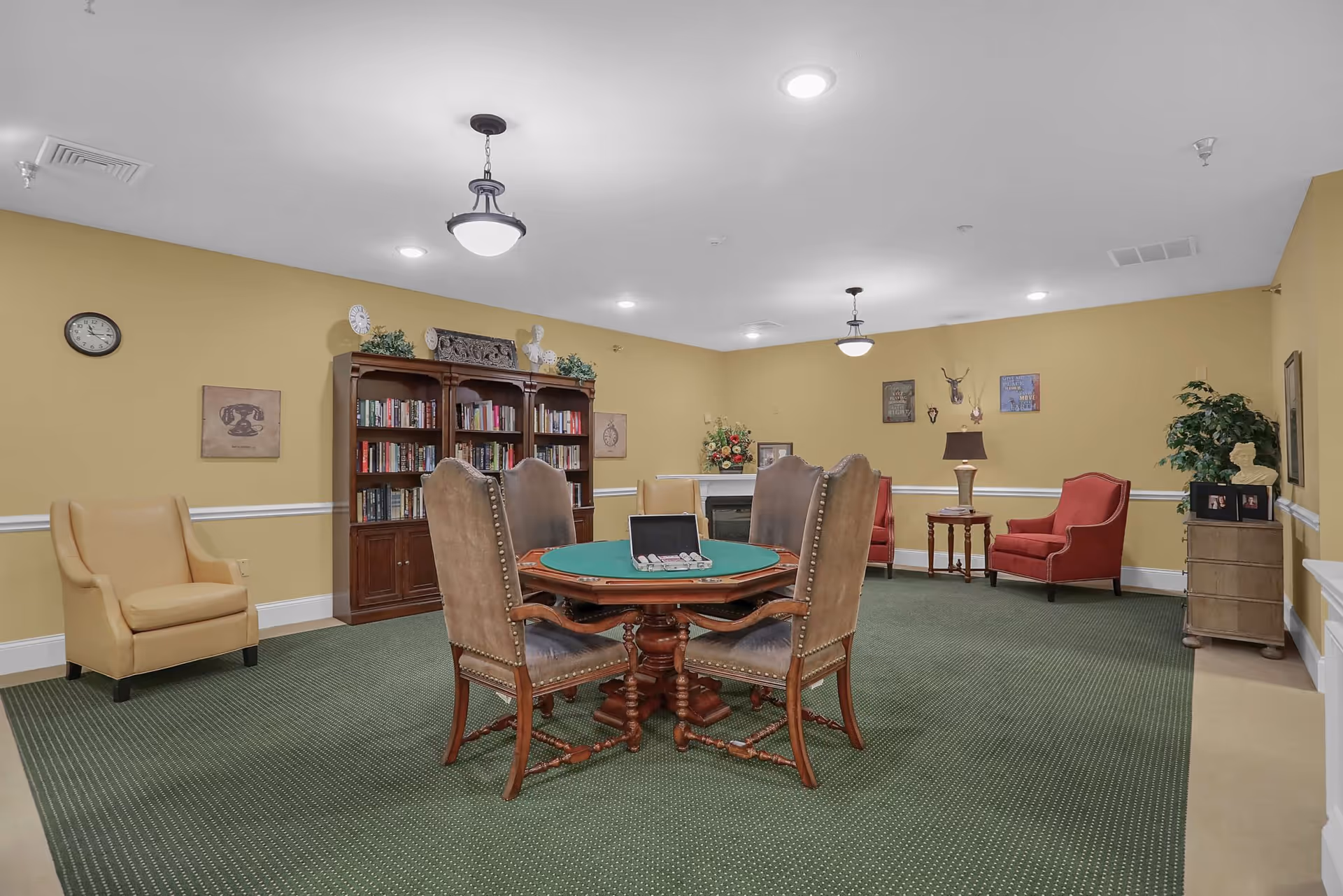 A cozy common room with yellow walls and green carpet featuring a round wooden table with four upholstered chairs in the center. A bookshelf filled with books is against the back wall, flanked by decorative items and a clock. There are two red armchairs with a small wooden table and lamp between them, and a beige armchair on the left side. The room is softly lit by ceiling lights and hanging fixtures.