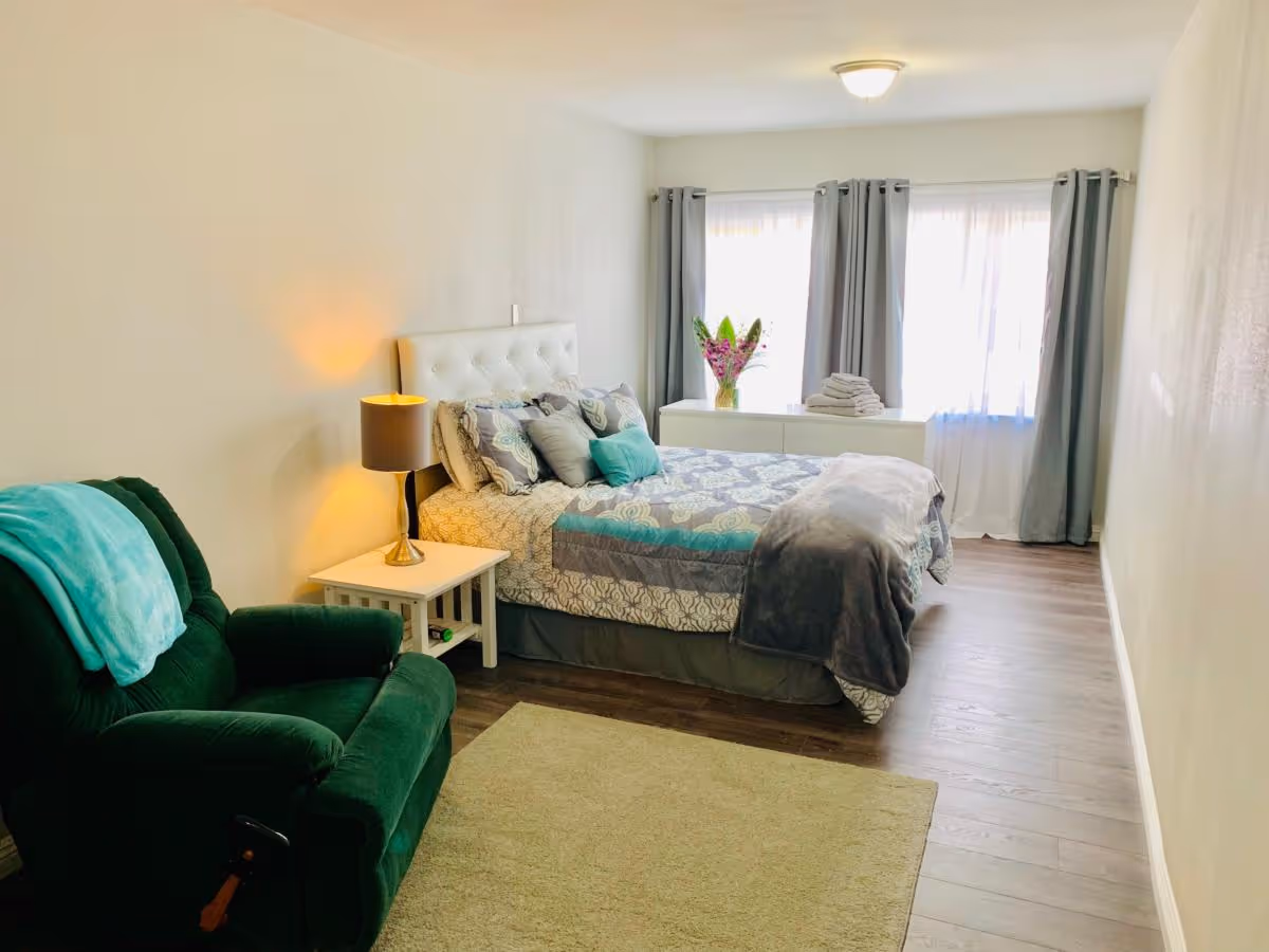 A bright bedroom with a bed featuring patterned bedding and multiple pillows, a white nightstand with a lamp, a green armchair with a teal blanket draped over it, a light-colored rug on wooden flooring, and a white dresser with a vase of flowers and folded towels in front of two large windows with gray curtains.