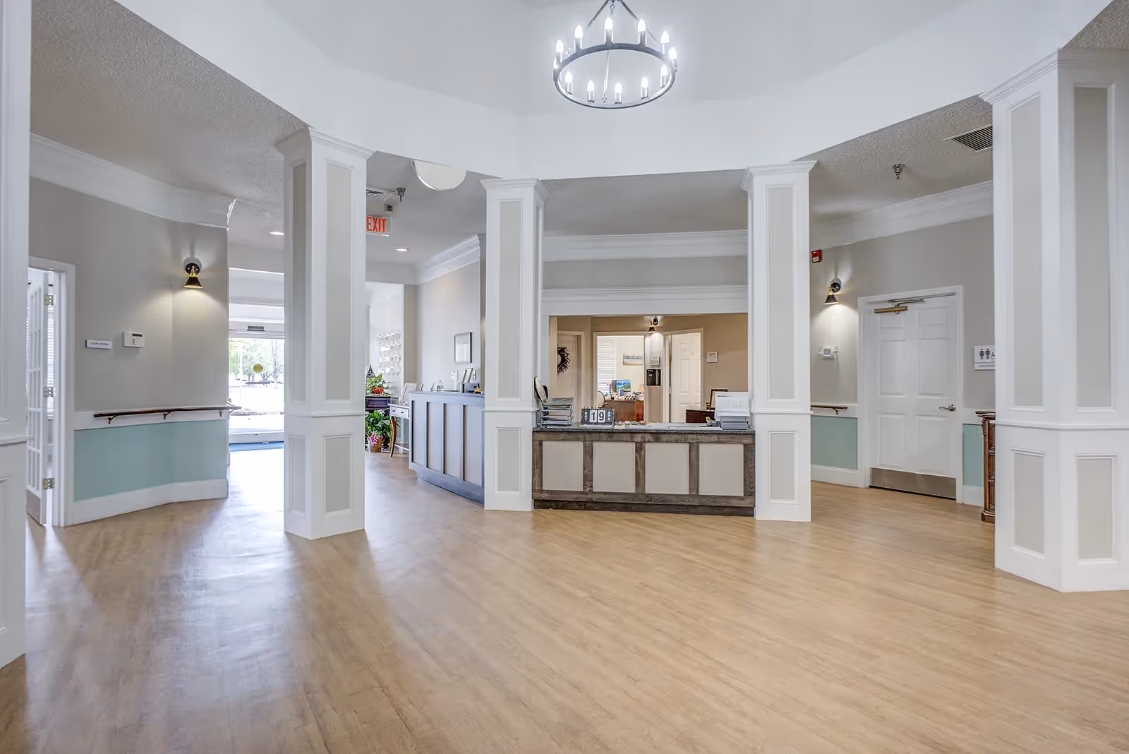 Spacious and well-lit reception area with light wood flooring, white columns, and a chandelier hanging from the ceiling. There is a reception desk in the center with some office equipment and a sign displaying the number 119. The walls are painted in neutral tones with white trim, and there are doorways leading to other rooms. The area appears clean and welcoming.