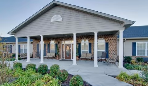 Front exterior view of a senior living facility with a covered entrance supported by white columns, brick walls, and a glass front door. There are benches on the porch and landscaped greenery in front.