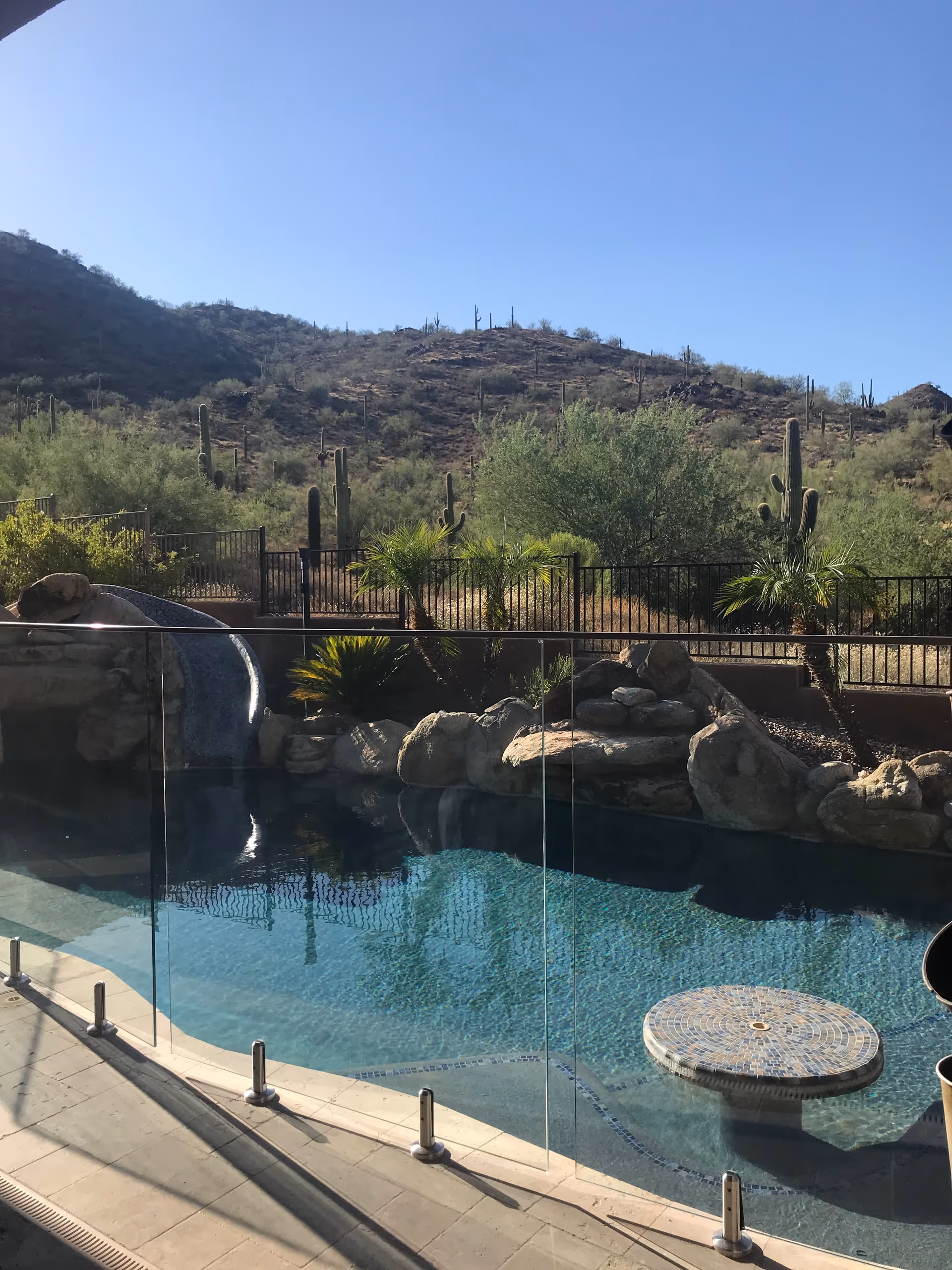 Outdoor swimming pool with a built-in round mosaic table in the water, surrounded by a glass safety fence. In the background, there are desert hills with cacti and sparse vegetation under a clear blue sky.