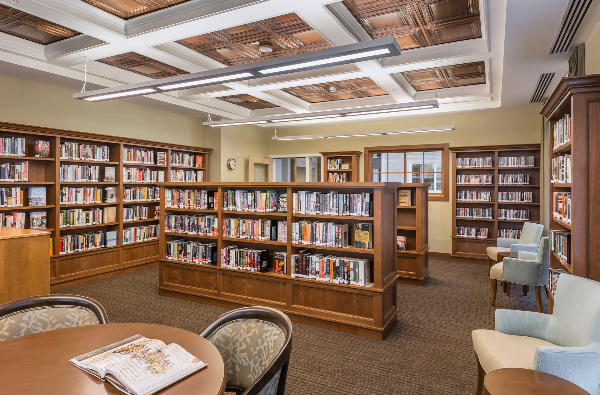 A well-lit library room with wooden bookshelves filled with books, a round table with an open book, and several upholstered chairs. The ceiling has decorative wooden panels and modern hanging lights.