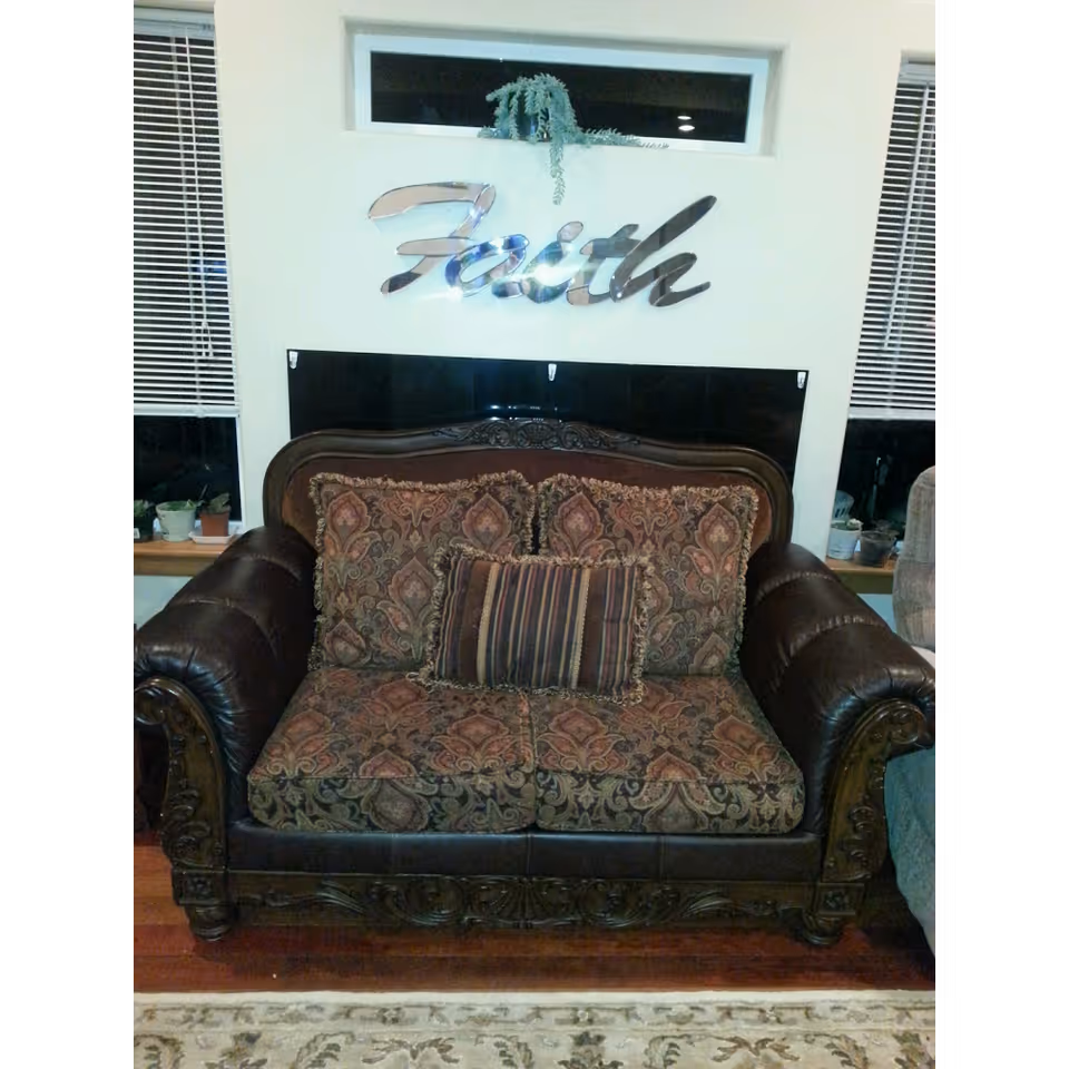 A vintage-style loveseat with dark leather armrests and ornate wooden carvings, upholstered with patterned fabric cushions. Above the loveseat, a decorative silver wall sign spells out the word 'Faith'. Two windows with blinds flank the loveseat, and small potted plants are placed on the window sills.