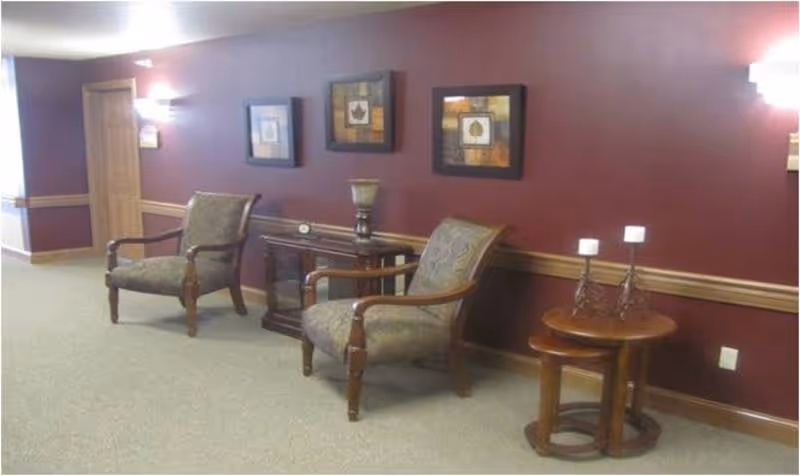 Two upholstered armchairs with side tables and framed artwork along a burgundy hallway wall in a common sitting area.