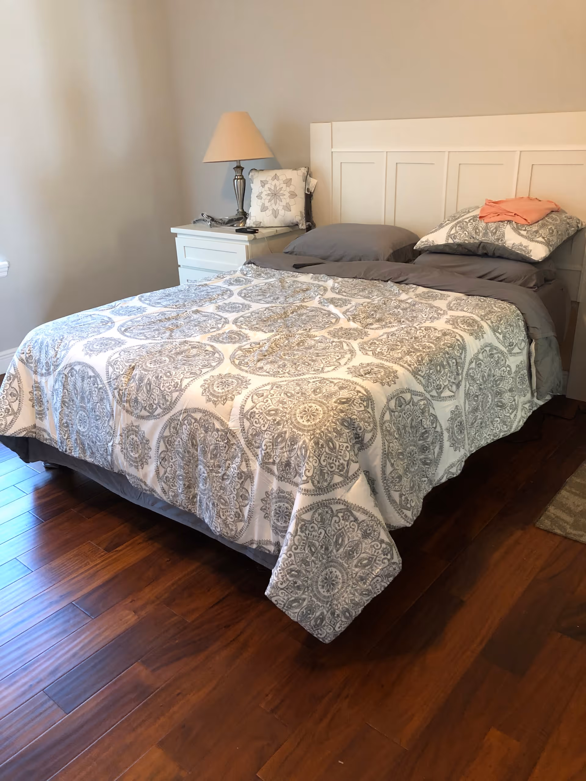 A neatly made bed with a white and gray patterned bedspread and gray pillows in a bedroom with wooden flooring. There is a white nightstand next to the bed with a lamp and a decorative pillow on it.